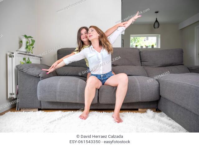 Playful and excited little girl holding her mother hands while pretending to fly for fun at home. Happy caucasian mother bonding and relaxing with her daughter
