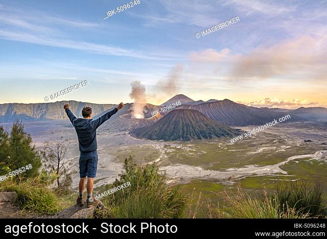 Young man stretches arms in the air, volcanic landscape at sunset, view in Tengger Caldera, smoking volcano Gunung Bromo, in front Mt