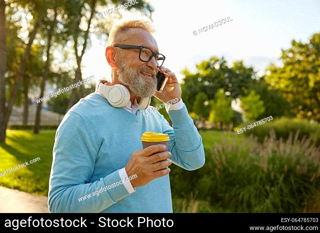Side view portrait of senior man having call on smartphone. Elderly businessman walking in park enjoying coffee break and talking with relatives on cellphone