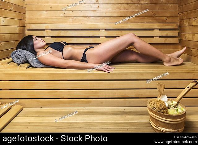 Side view of beautiful slim woman in black bikini lying on wooden shelf in sauna