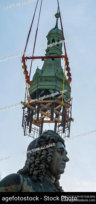11 November 2021, Saxony-Anhalt, Halle (Saale): The tower helmet of the Halle/Saale town house hovers on the crane hook behind the Handel monument from the...