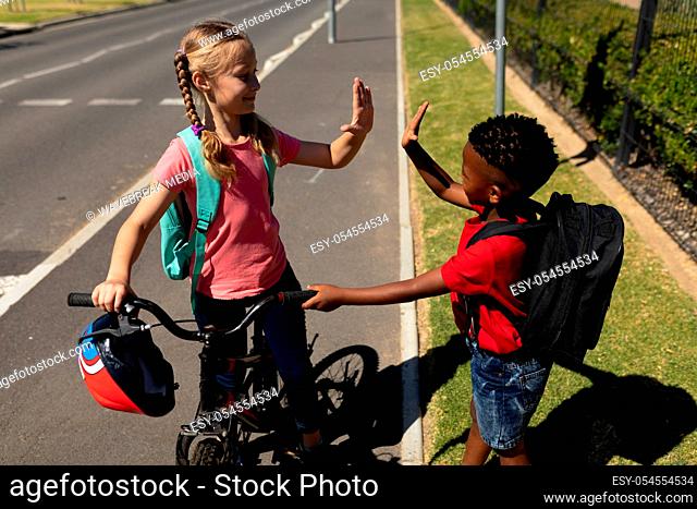 Side view of a Caucasian schoolgirl with plaits and an African American schoolboy wearing a red t shirt standing on the pavement