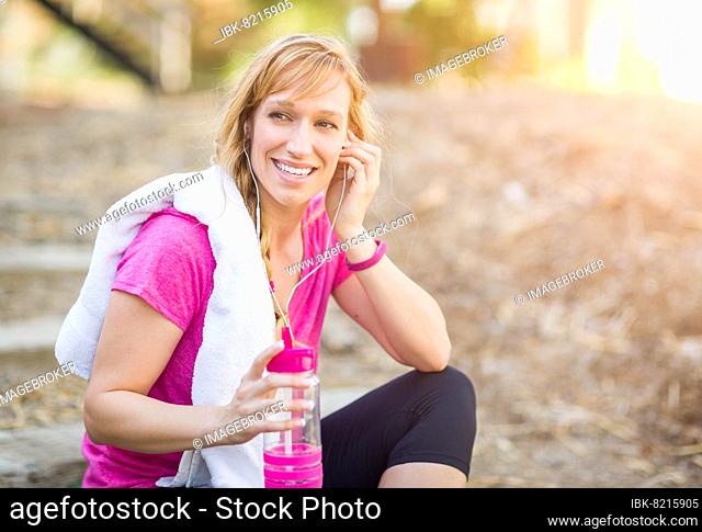 Young fit adult woman outdoors with towel and water bottle in workout clothes listening to music with earphones