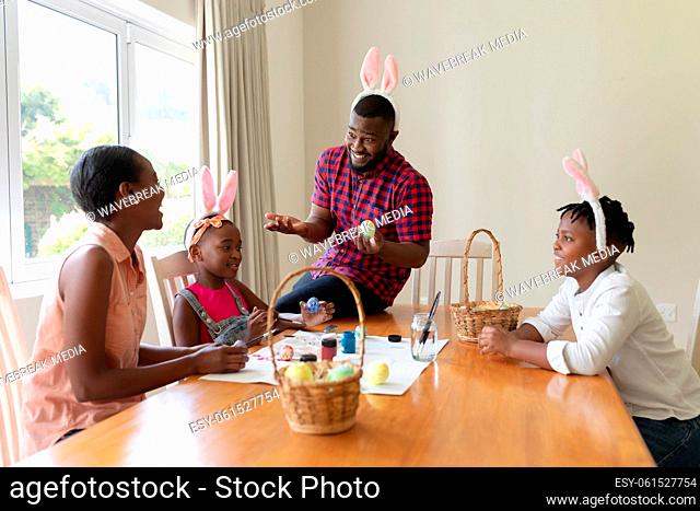 Happy african american parents with son and daughter wearing bunny ears painting easter eggs
