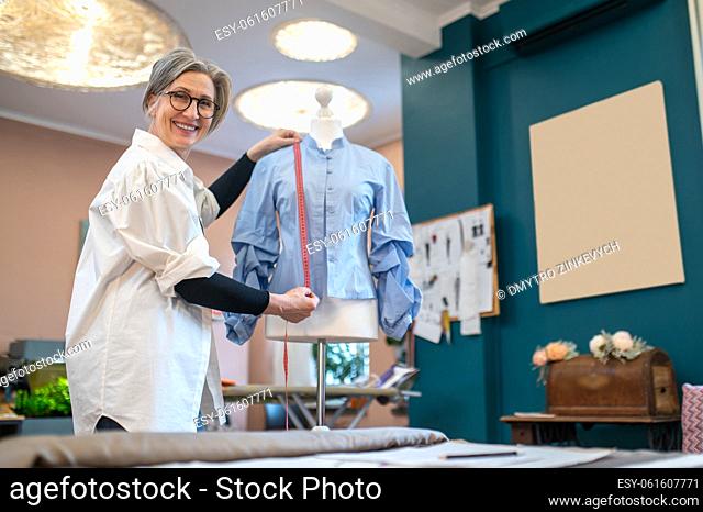 Good mood. Gray-haired friendly woman in glasses standing with measure near blouse on mannequin smiling at camera in atelier