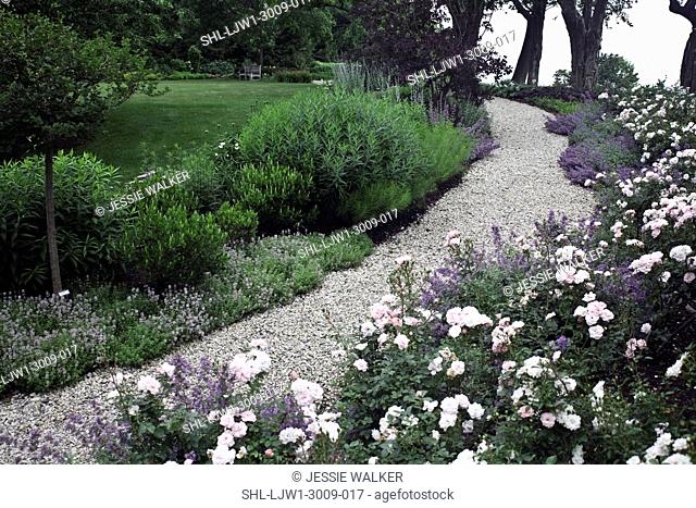 GARDENS: Curving gravle path, fairy roses in foreground, path is bordered by flowers, and treeline, lawn to left with more flower beds beyond