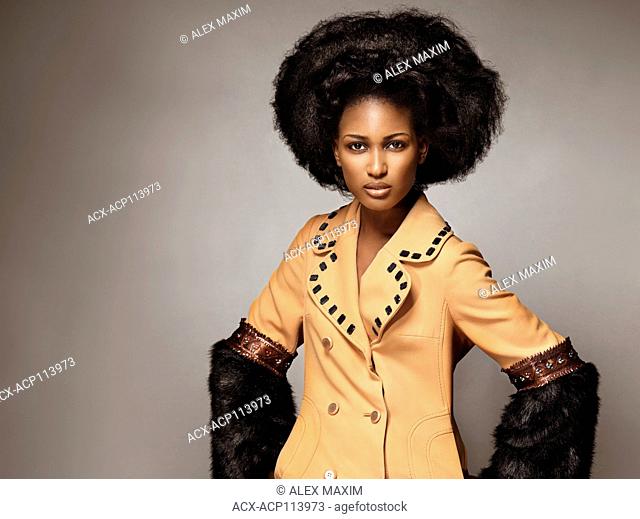 High fashion edgy beauty portrait of a black african american woman wearing an orange coat with black fur on gray background