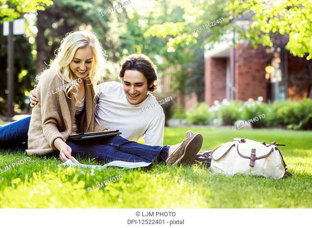 A young couple studying outdoors on the grass on the university campus and looking through a textbook while using a tablet; Edmonton, Alberta, Canada