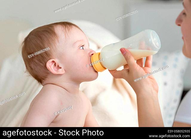 Mother giving milk from bottle to her baby son sitting on sofa