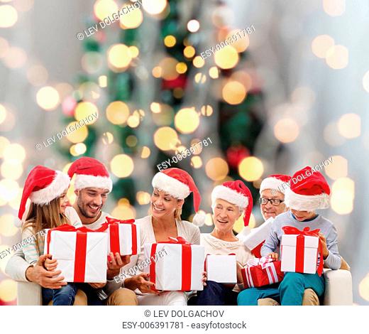 family, happiness, generation, holidays and people concept - happy family in santa helper hats with gift boxes sitting on couch over christmas tree lights...