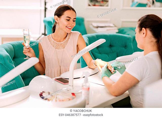 Champagne and manicure. Beaming elegant businesswoman drinking glass of champagne and getting her manicure