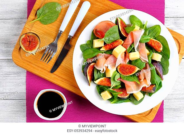 fresh baby spinach, figs, thinly sliced italian ham and tender mozzarella cheese salad on white plate on cutting board with dressing and cutlery on table mat on...