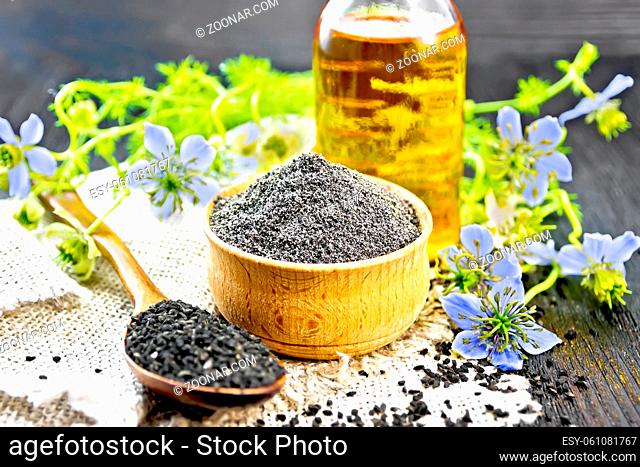 Flour of black caraway in a bowl, seeds in a spoon burlap napkin, oil in bottle and twigs Nigella sativa with blue flowers and leaves on dark wooden board...