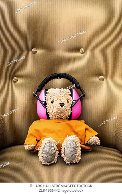Conceptual image of teddybear wearing orange sweater and pink hearing protection sitting in armchair