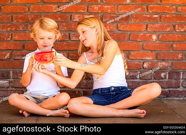 Cute Young Cuacasian Boy and Girl Eating Watermelon Against Brick Wall