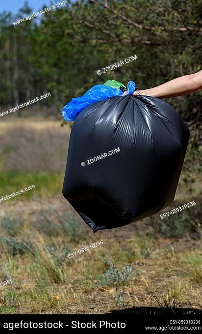 hand holds against a forest full of garbage a large black plastic bag, a summer day, garbage collection in nature