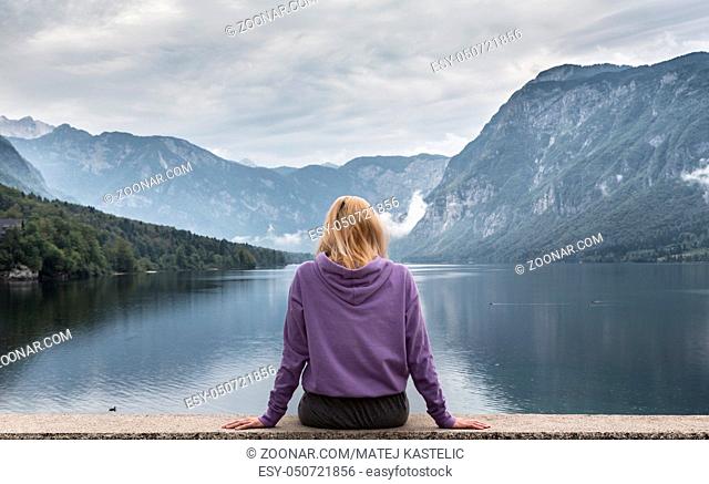 Solitary woman wearing purple hoodie watching tranquil overcast morning scene at lake Bohinj, Alps mountains, Slovenia