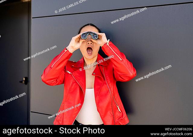 Shocked woman with mouth open looking through binoculars in front of gray wall
