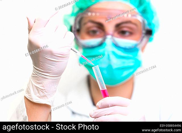 Young attractive concentrated female scientist in protective eyeglasses, mask and gloves dropping a red liquid substance into the test tube with a pipette in...