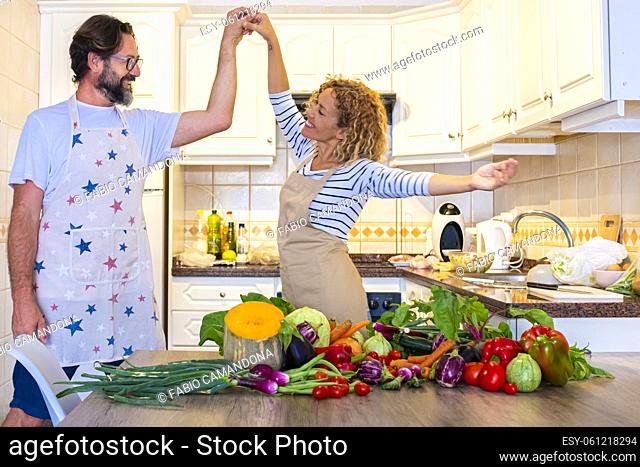 Happy adult couple dance and have fun together in the kitchen at home while preparing healthy vegetables on the table. Overjoyed woman and man in love prepare...