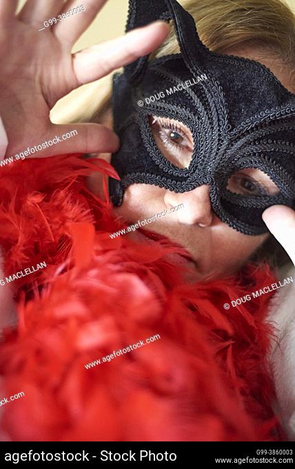 Portrait of a fifty plus years old woman with brown eyes wearing a textured black mask and red feather boa with her hand by her face