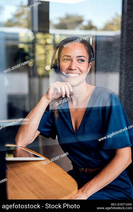 Smiling thoughtful businesswoman wearing headset seen through glass