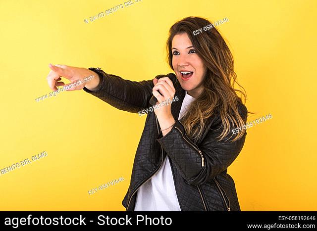 Young singer girl, wearing black leather jacket, singing with microphone and pointing with her hand on yellow background