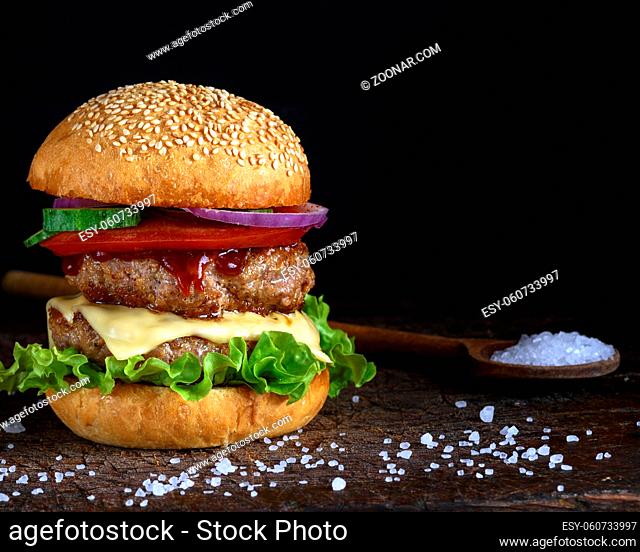 cheeseburger in a bun with sesame seeds, in the middle fresh vegetables and meat cutlets, black background
