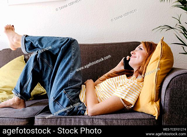 Young woman talking on smart phone while lying on sofa at home