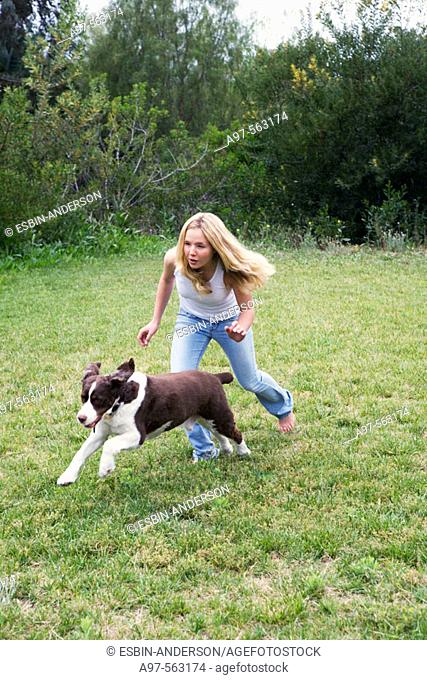 Barefoot blonde teen girl in blue jeans and white tank top running with pet dog on a grass lawn