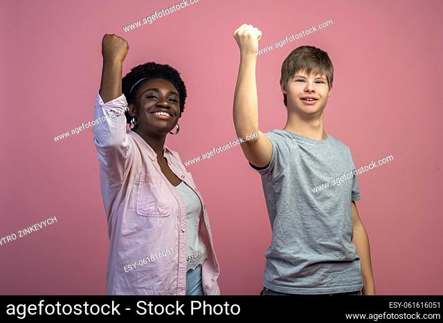 Team. African american woman and caucasian guy with down syndrome with raised fist looking at camera standing on light background