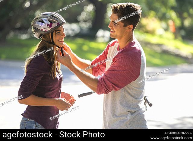 Young couple getting ready for bike ride, man fastening the strap on the woman helmet