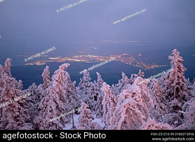 16 December 2023, Saxony-Anhalt, Schierke: View over snow-covered spruce trees to the lights of the town of Wernigerode. The mild temperatures of the past few...
