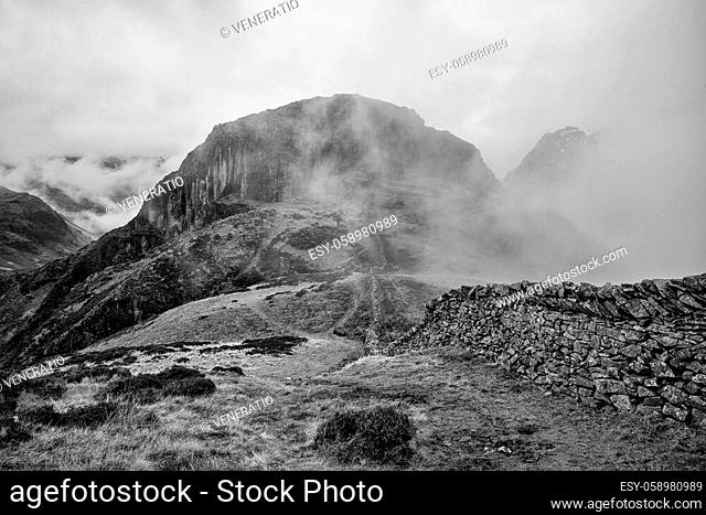 Stunning black and white Winter landscape image of view from Side Pike towards Langdale pikes with low level clouds on mountain tops and moody mist
