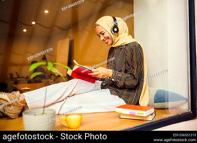 Beautiful arab girl in hijab and headphones in university cafe. Muslim woman with books sitting in library. Religion and education
