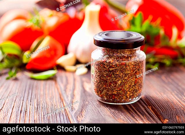 Closeup of the small jar with dried chopped spices located on a wooden table in the rustic style. The fresh vegetables on the background
