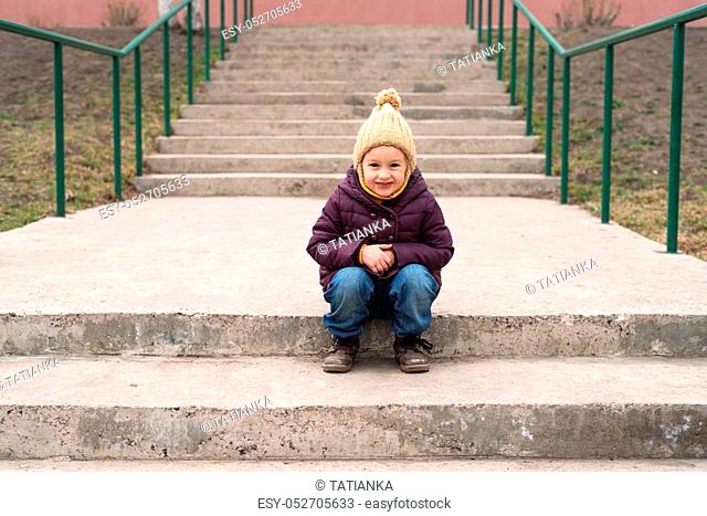 Little girl in a violet coat and a beige woolen hat sitting on the stairs and smilling
