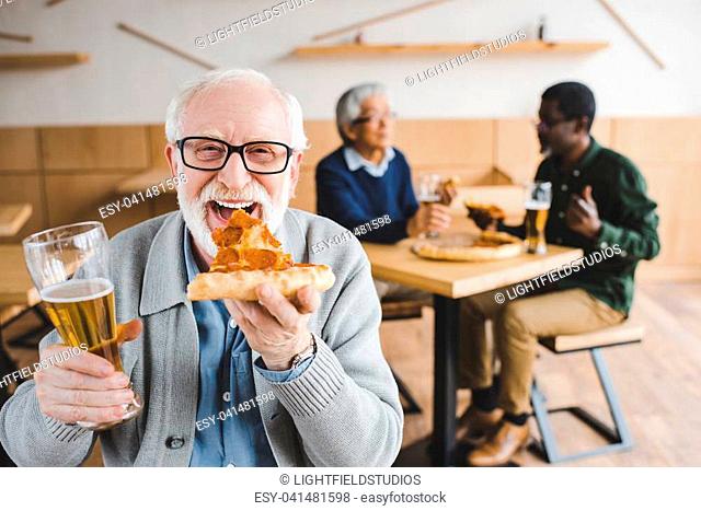 happy senior man eating pizza with beer while his friends talking on background