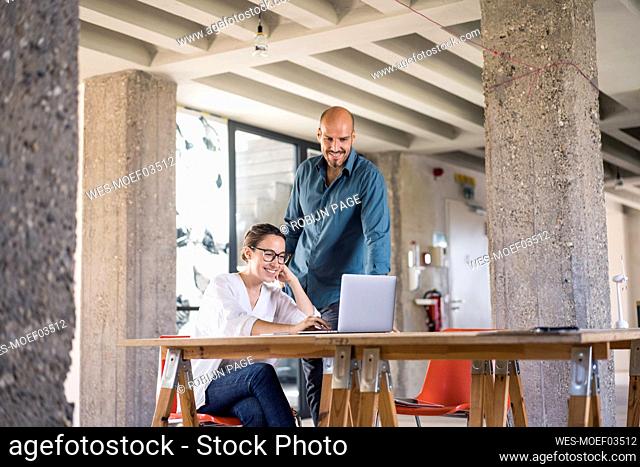 Smiling woman using laptop by man standing at table in office