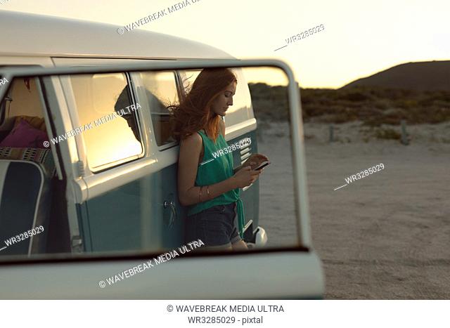 Side view of beautiful Caucasian woman leaning on camper van and using mobile phone at beach