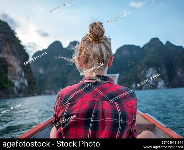 Back view of woman sitting in wood long tail boat over tropical limestone cliffs and sunset background