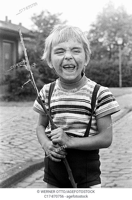 Seventies, black and white photo, people, children, little boy, portrait, street kid, dirty, unkemptly, holds a stick in his hands and makes a face