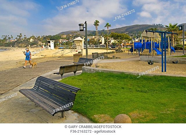 Woman walking her dog holding coffee on boardwalk next to sand at Laguna Beach, Orange County, California