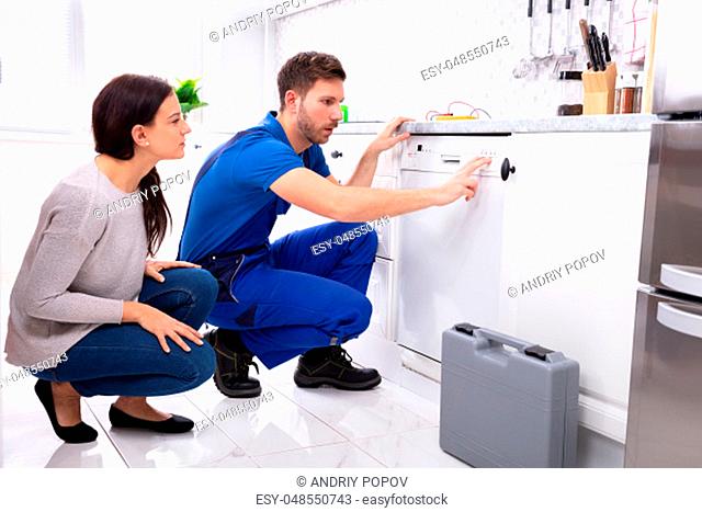 Serviceman Crouching On Kitchen Floor Pressing Button Of Dishwasher While Woman Looking At Him