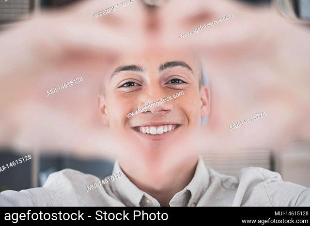 Cheerful affectionate teenager man take romantic selfie portrait looking at camera through heart of joined fingers. Enamored young lady shoot love confession to...