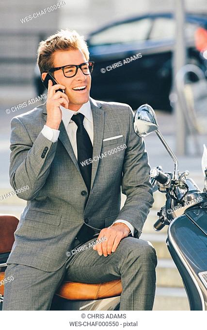 Smiling businessman sitting on motor scooter talking on cell phone