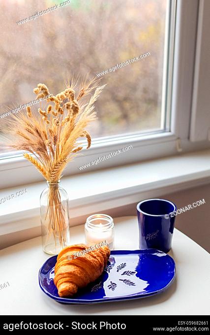 Handmade blue clay plate with croissant on it. Vase with wheat and candles