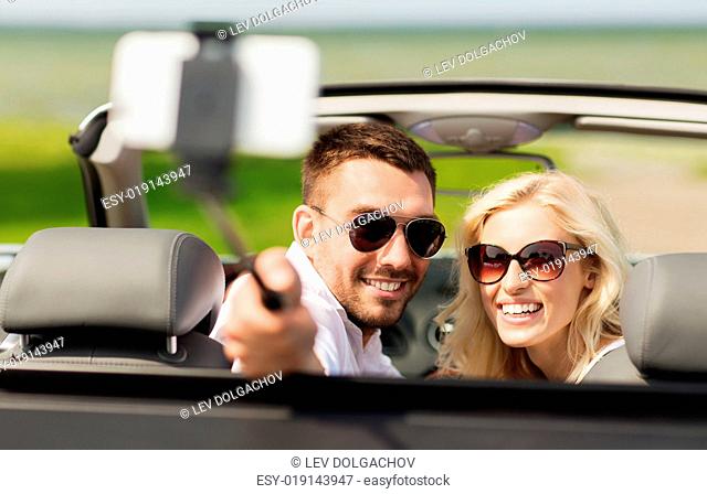 road trip, leisure, couple, technology and people concept - happy man and woman driving in cabriolet car and taking picture with smartphone on selfie stick