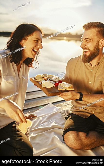Happy couple having picnic on jetty at a lake at sunset eating cheese