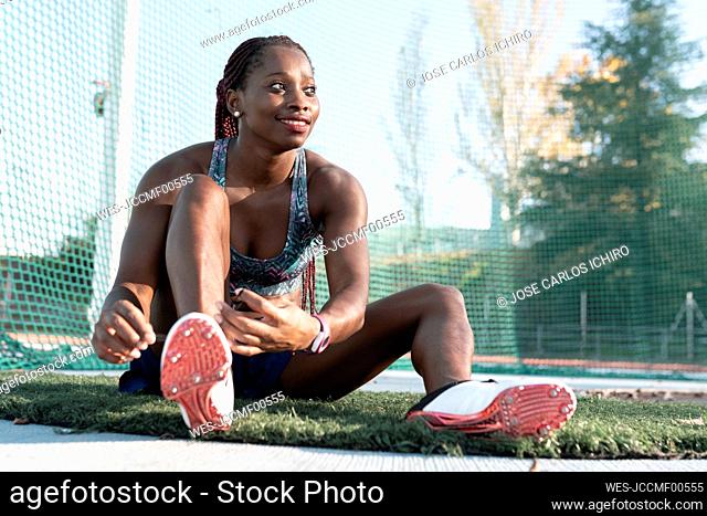 Smiling young sportswoman looking away while tying shoelace against net